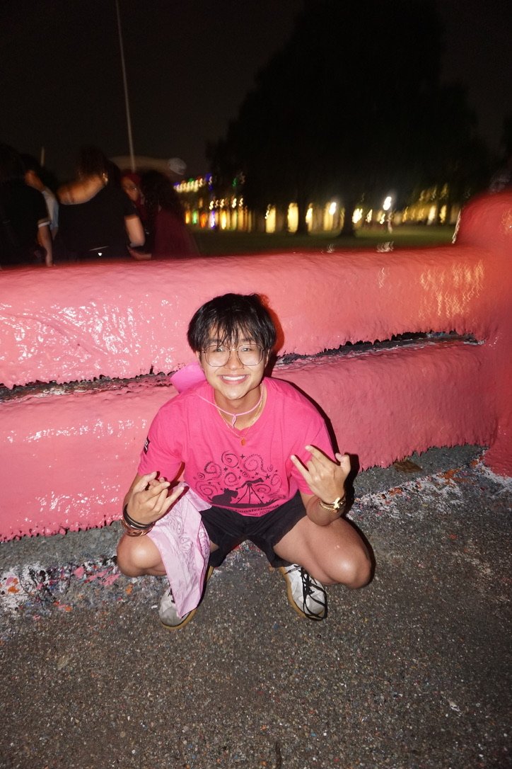 Jin crouching in a pink shirt in front of a pink-painted fence at night