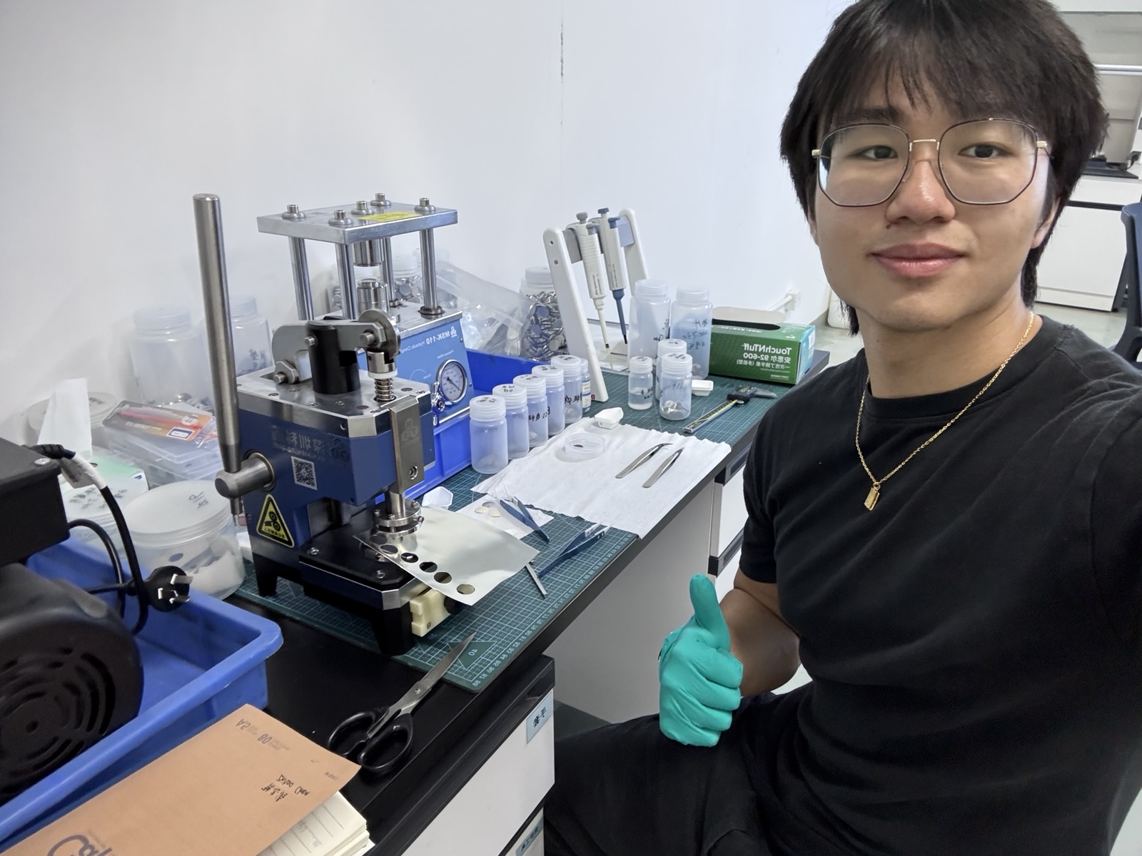 Jin giving a thumbs up beside the coin-cell press and electrode punch in the R&D lab